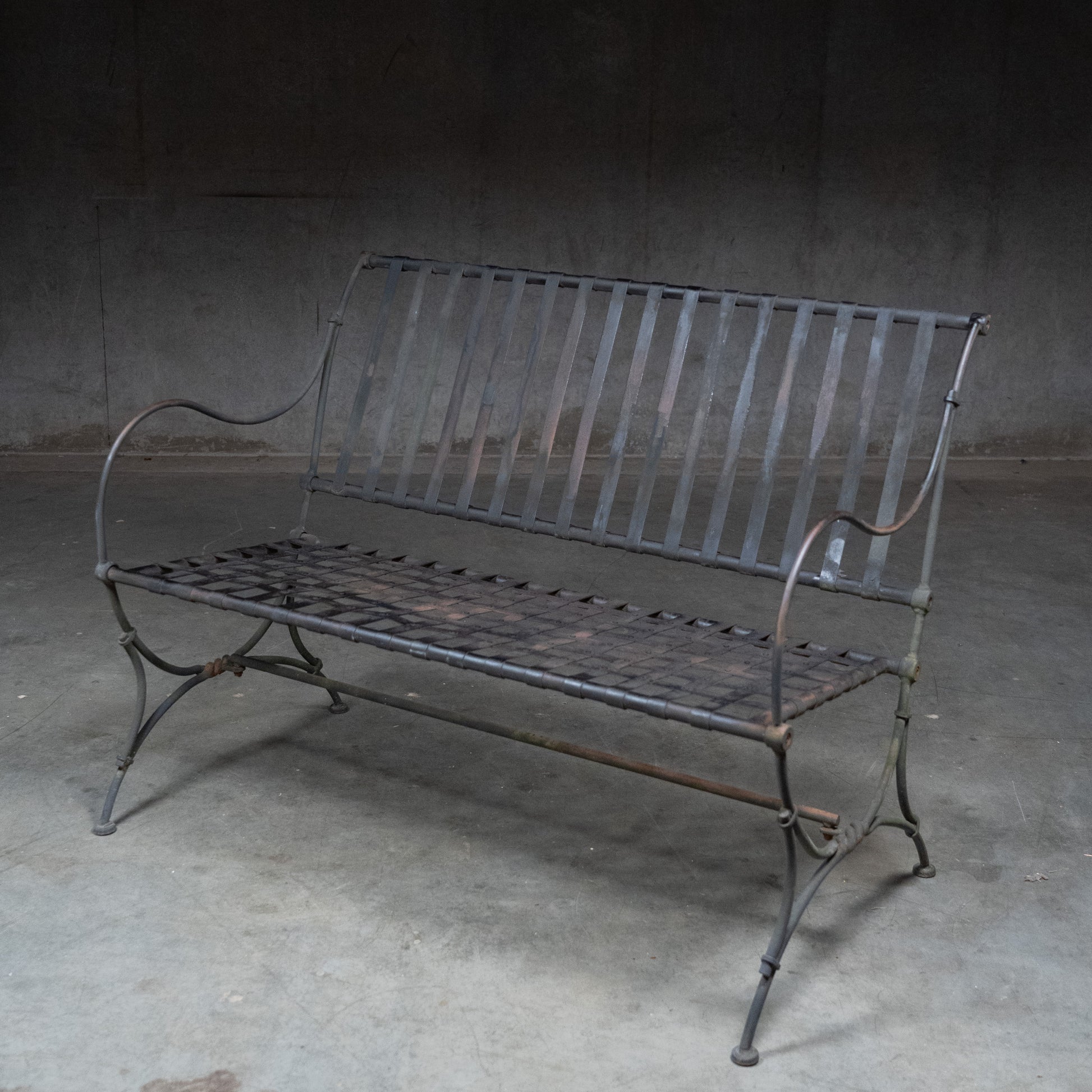 Vintage 1960s wrought iron garden bench with woven strap seat and back, curved arms and weathered painted finish./ Scott Landon Antiques and Interiors.